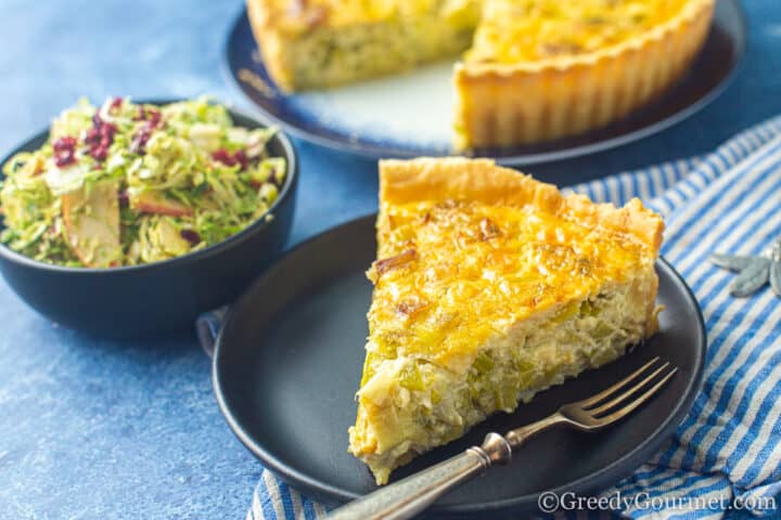 Slice of Leek tart on a plate with a bowl of salad.