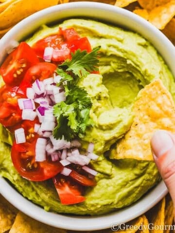 a bowl of creamy avocado dip, topped with chopped tomatoes, onions and cilantro, surrounded by tortilla chips