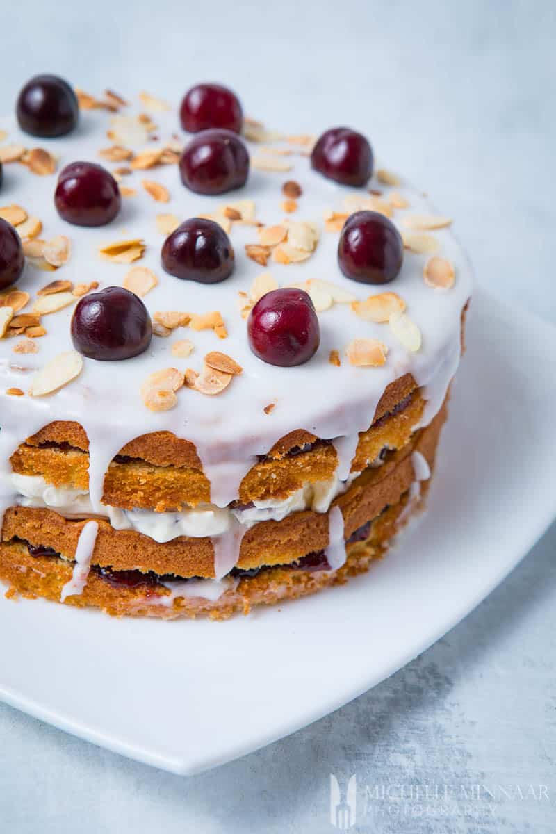 A finished cherry bakewell cake - four layers of cake with frosting drizzled on the sides and cherries on top 