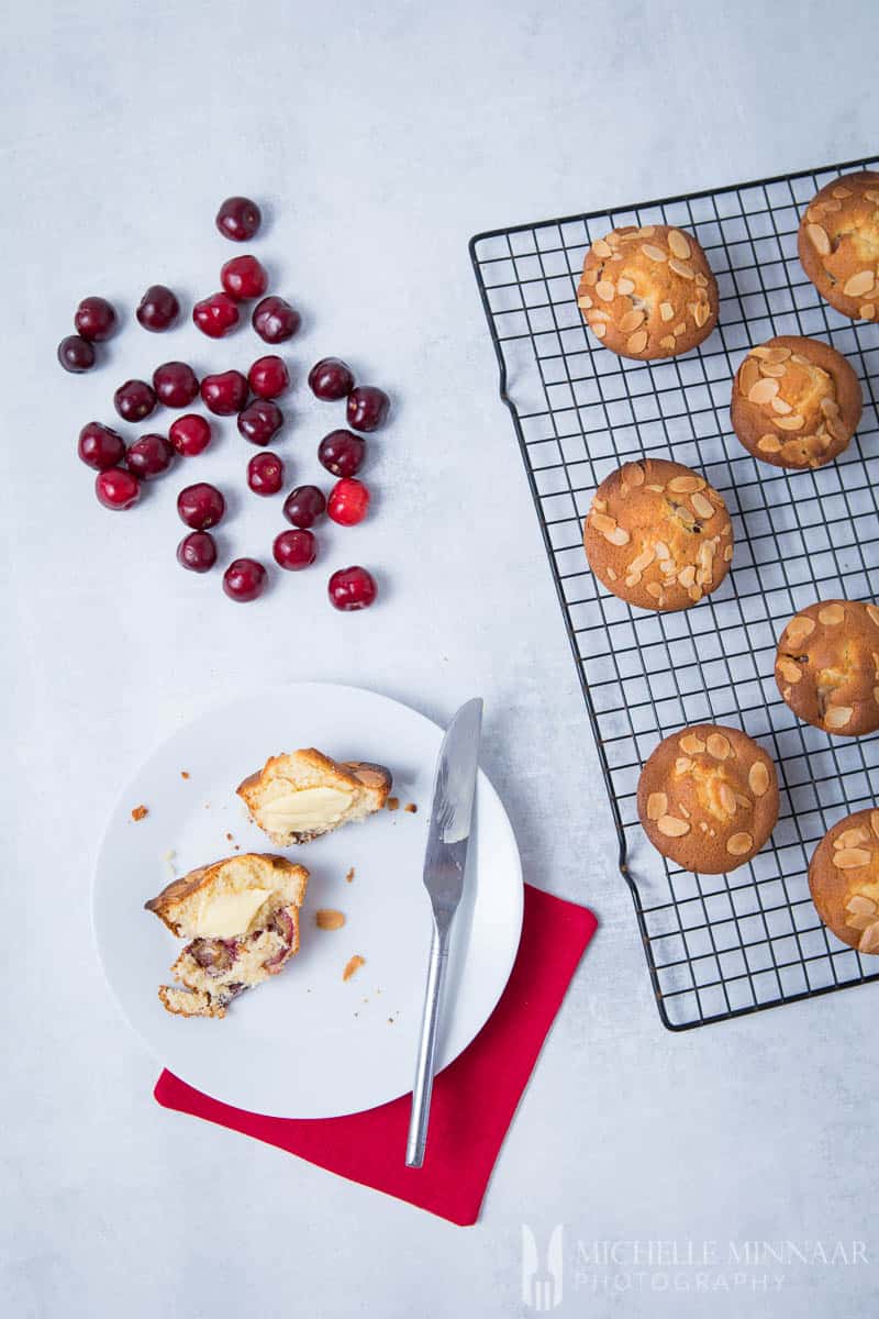 Loose cherries, muffins on a wire rack and a plate with one cherry muffin cut in half 