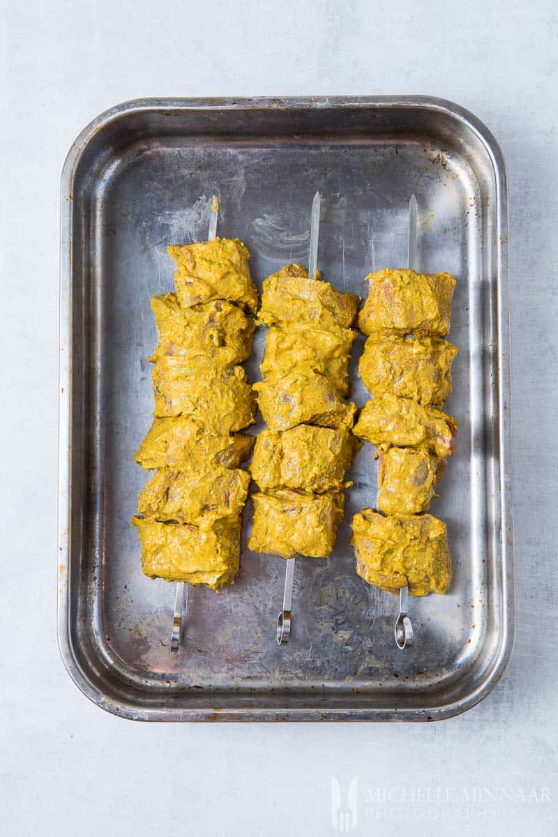 Raw Lamb covered in a yellow tikka sauce on skewers in a metal pan