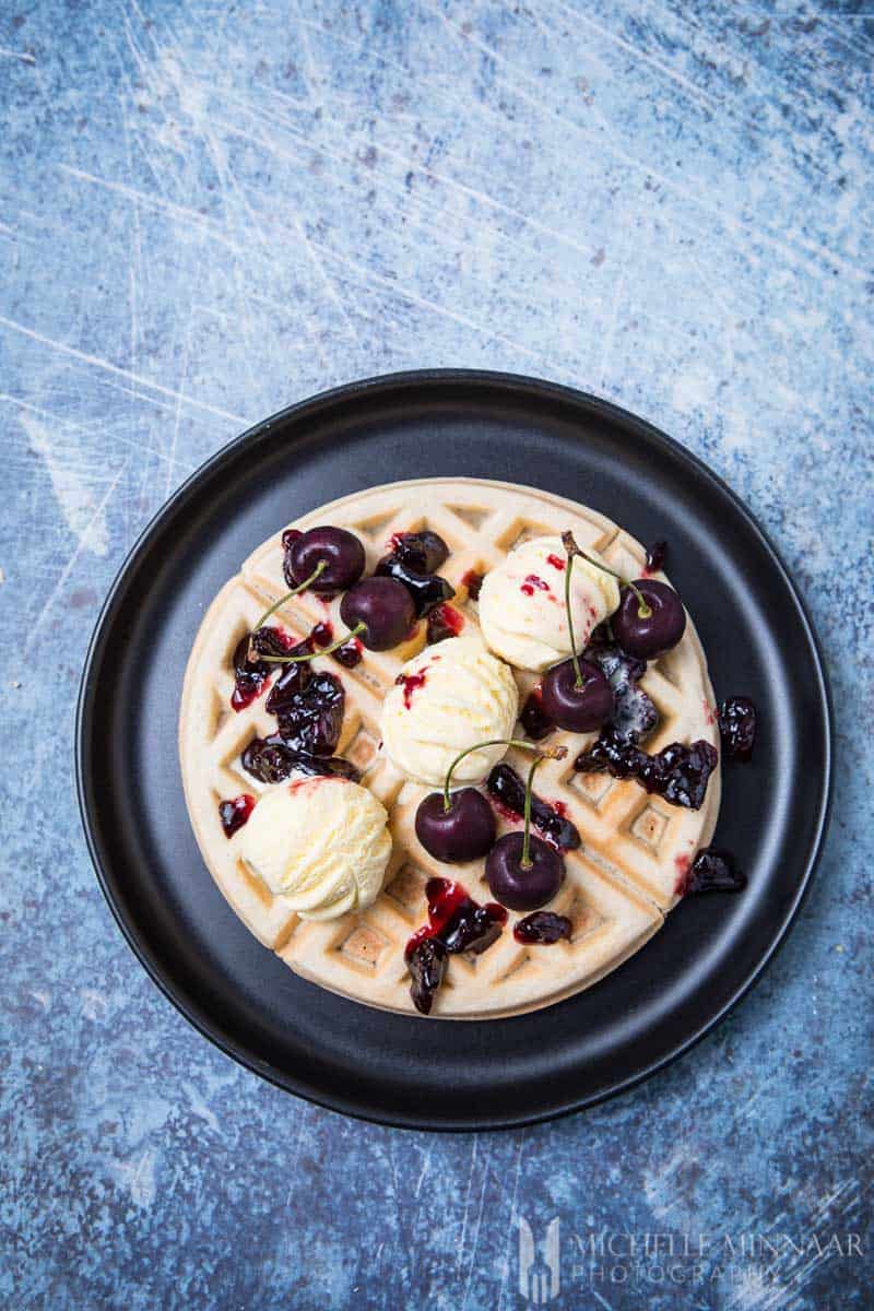 A dairy free waffles on a black plate with ice cream and cherries on top 