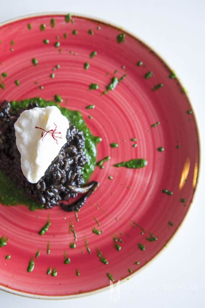 A pink plate with black risotto with a white dollap of sauce on top 