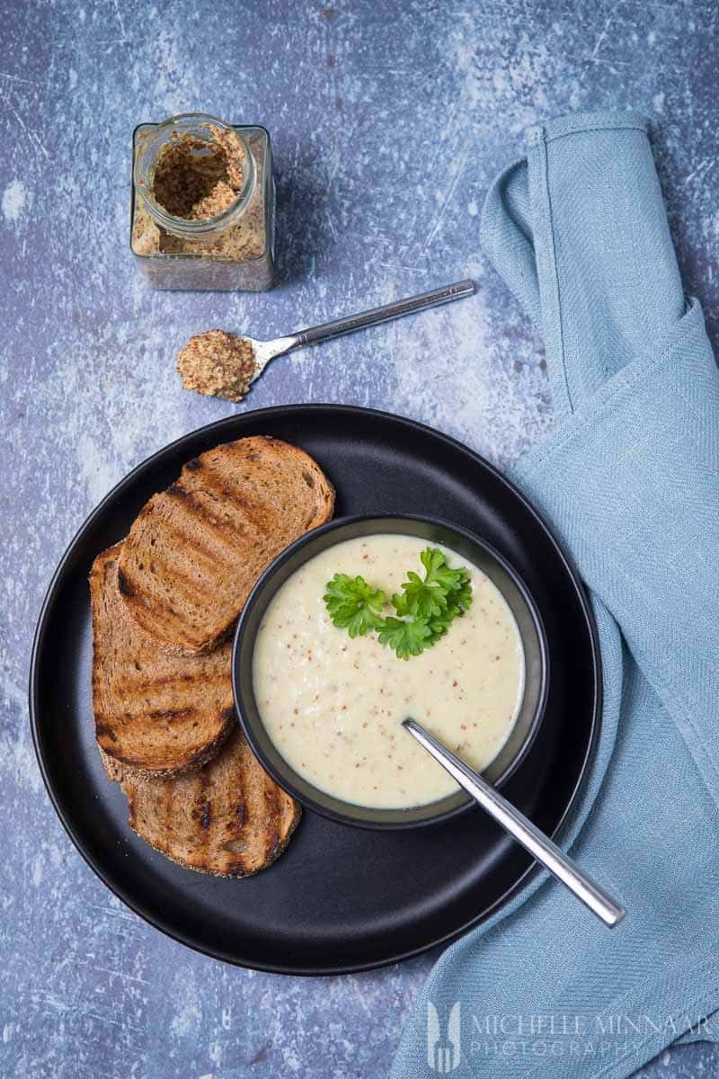 A bowl of mustard soup with bread on the side and a spoon with mustard on it 