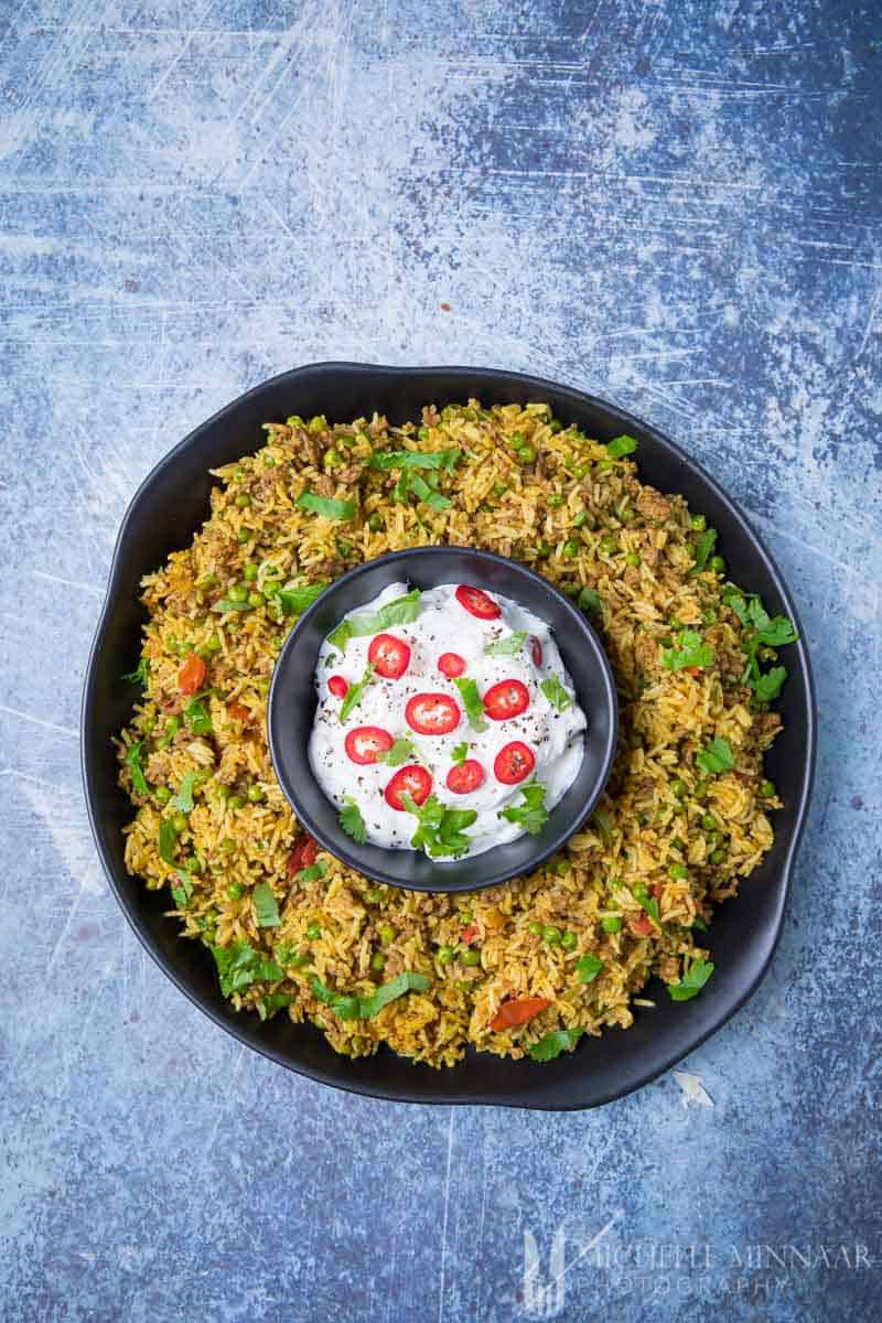 A large bowl of yellow keema rice with a bowl of yoghurt in the middle 