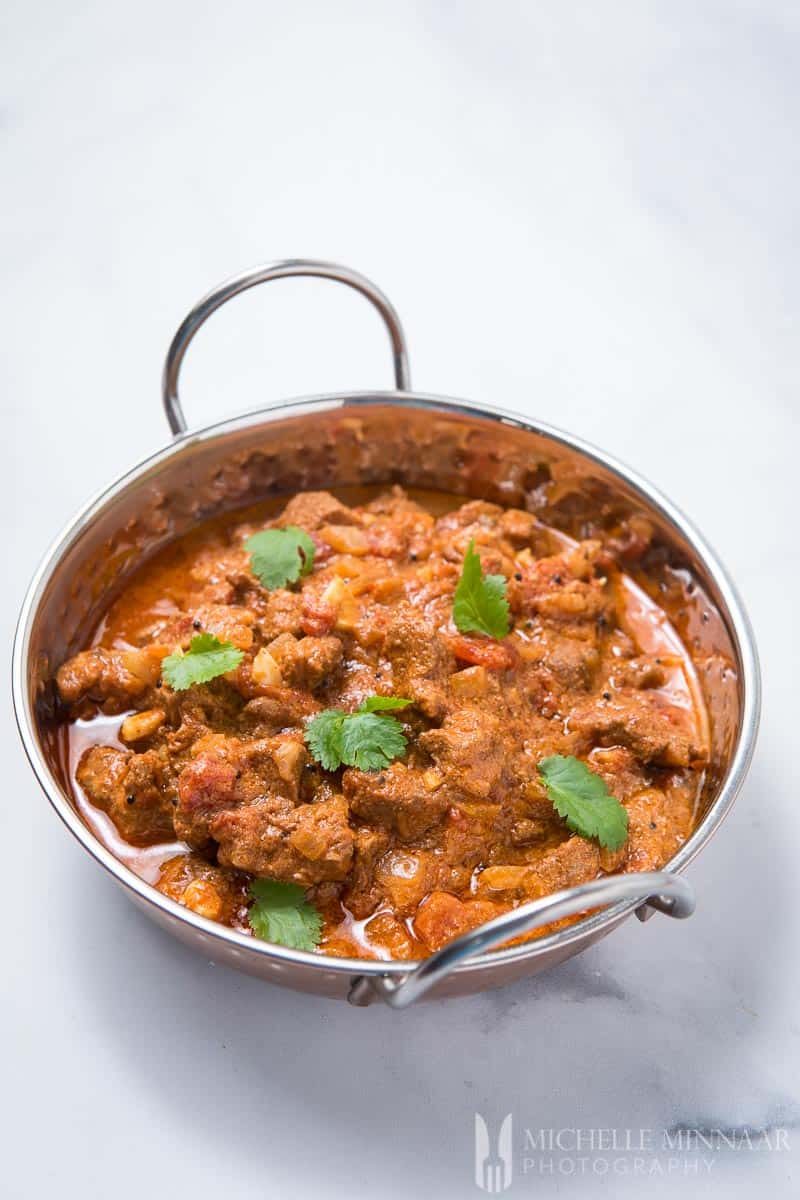 A bowl of browned lamb with a brown sauce in a silver pan for Lamb Madras