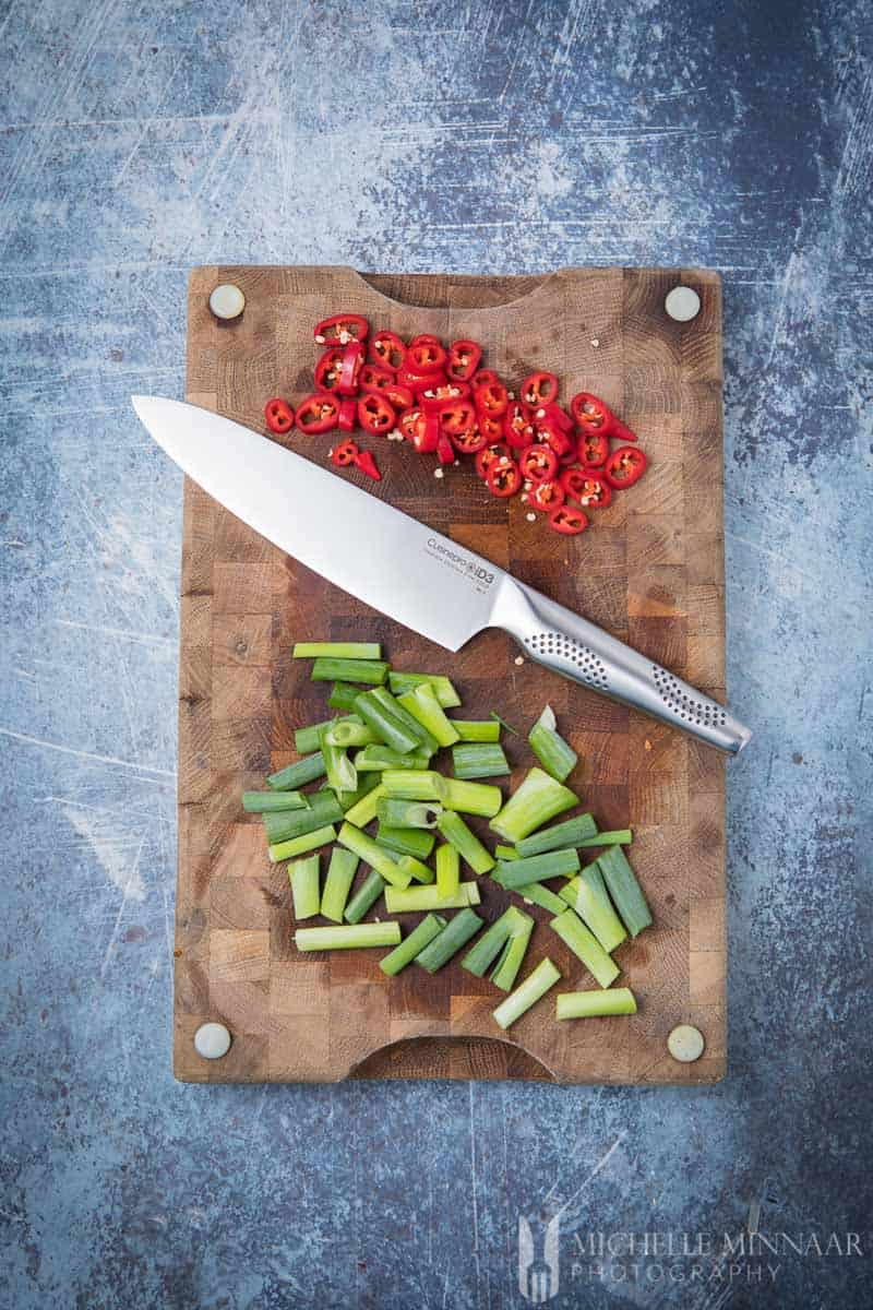 A large knife on a cutting board with Spring Onions and Chillies chopped up 