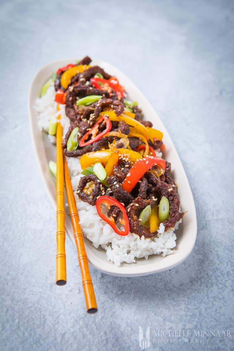 A plate of crispy chilli beef on top of white rice with chop sticks