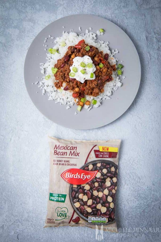 A plate of venison chilli on top of white rice with a bag of mexican bean mix next to it
