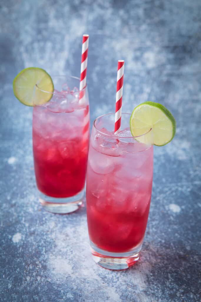 Two glasses of red and clear long beach iced tea with lime wheels as garnish and a red and white straw 