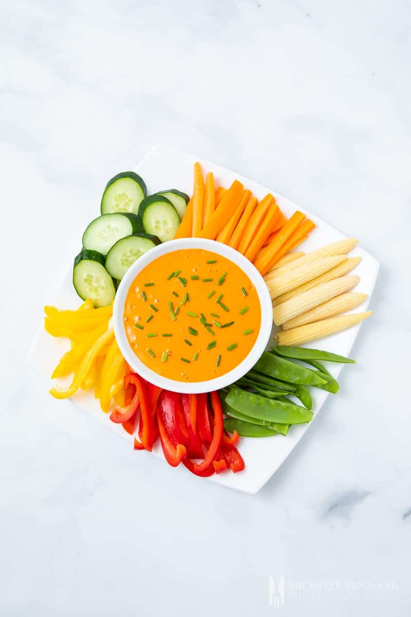 A bowl of orange harissa aioli with colorful cut vegetables 