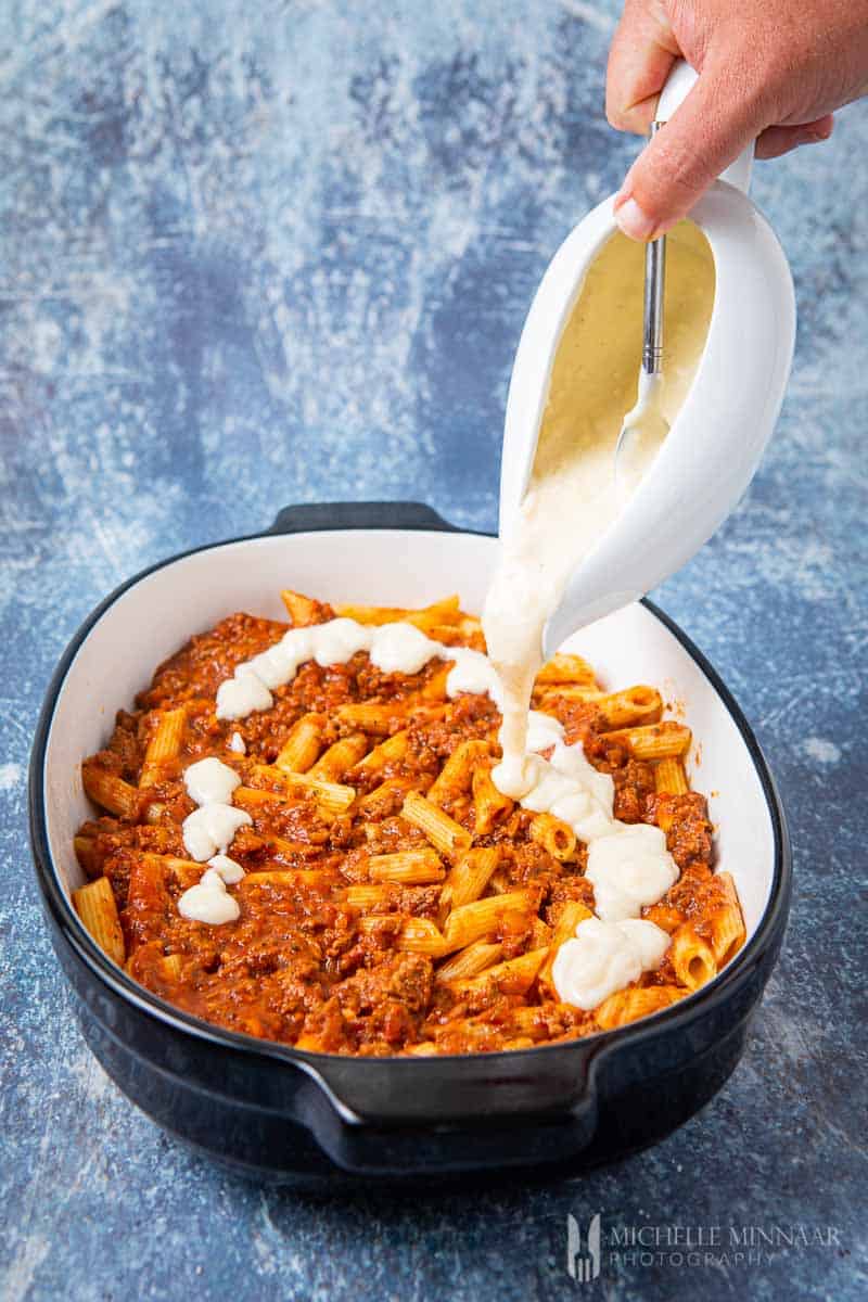 White sauce being poured on top of bolognese pasta bake