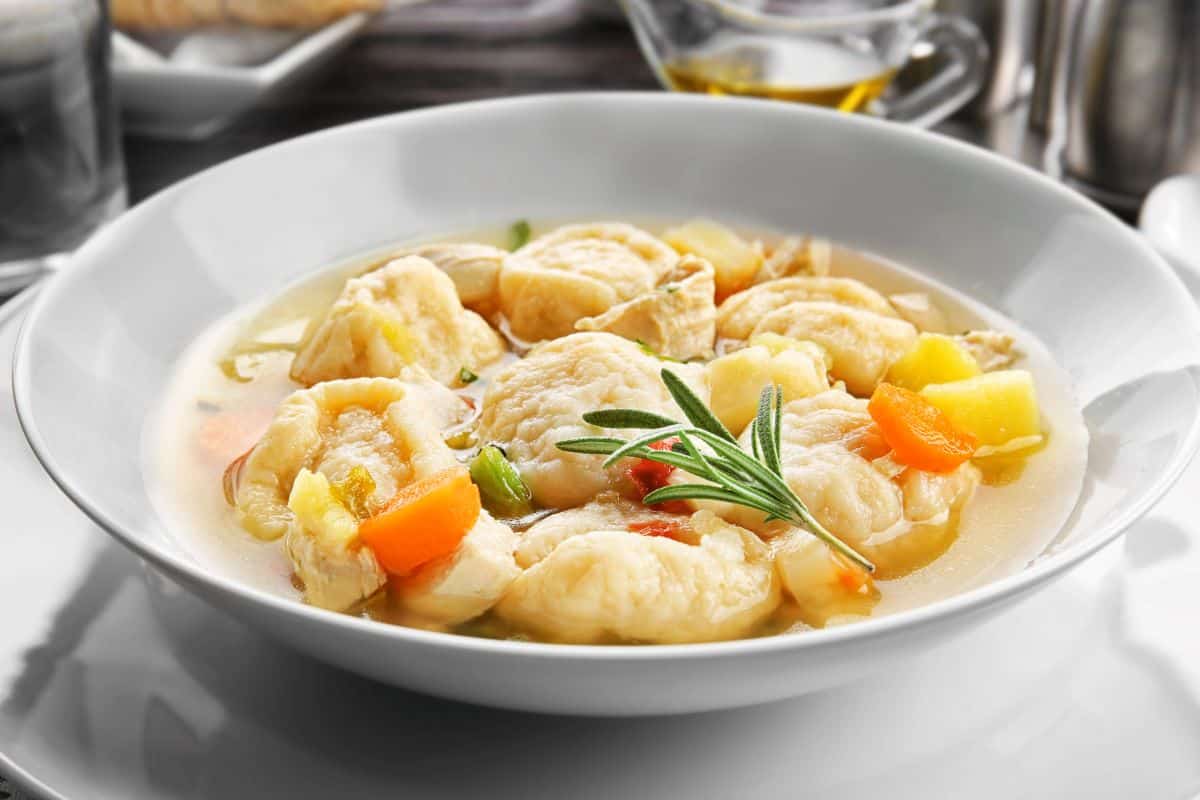 Portion of chicken and dumplings in a white bowl with vegetable pieces and herbs.