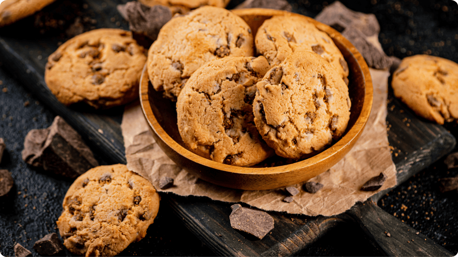 Chocolate chip cookies are arranged in a wooden bowl atop a cutting board, with scattered cookies and chocolate chips displayed on a black countertop.⁣