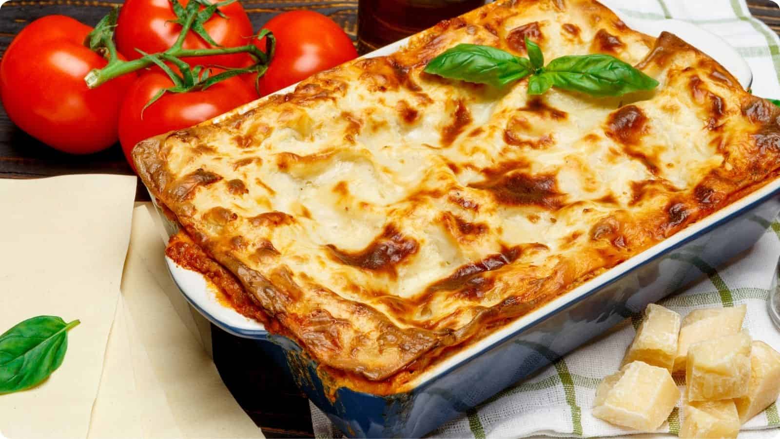 Freshly baked lasagna in a baking dish on a wooden table served with tomatoes and parmesan on the side on top of a wooden table.⁣
⁣⁣⁣⁣⁣⁣⁣⁣⁣⁣⁣⁣⁣
