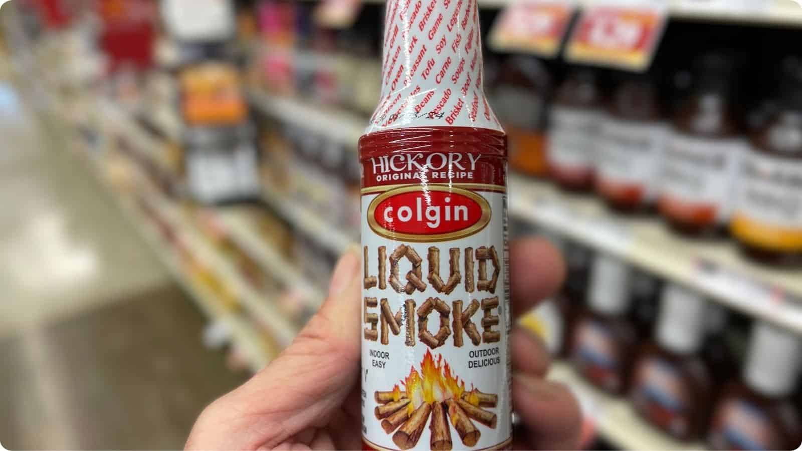 Close-up view of a hand holding a bottle of liquid smoke against a grocery store background.⁣