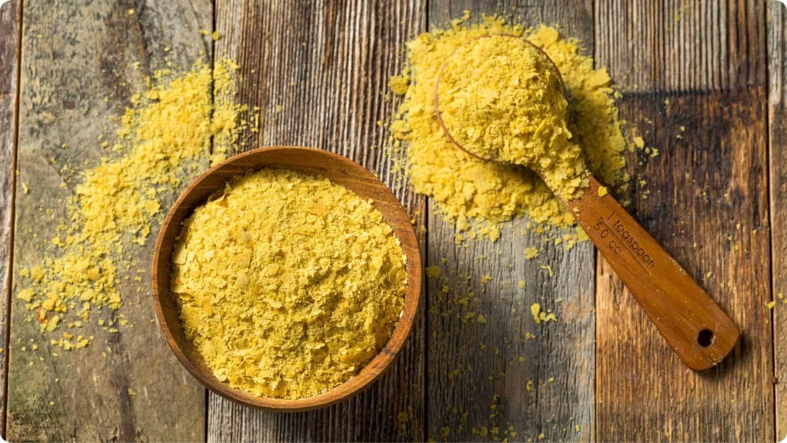 Overflowing bowl of yellow raw organic nutritional yeast with a scoop full on a wooden table.⁣⁣⁣⁣