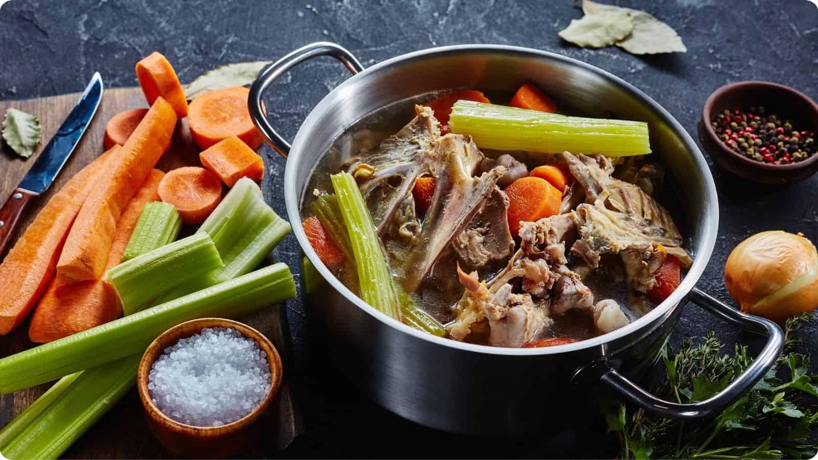 Cooked chicken stock surrounded by slices of carrots, onion, and sprigs of celery, placed on a charcoal-colored countertop.⁣
⁣⁣⁣⁣⁣⁣⁣