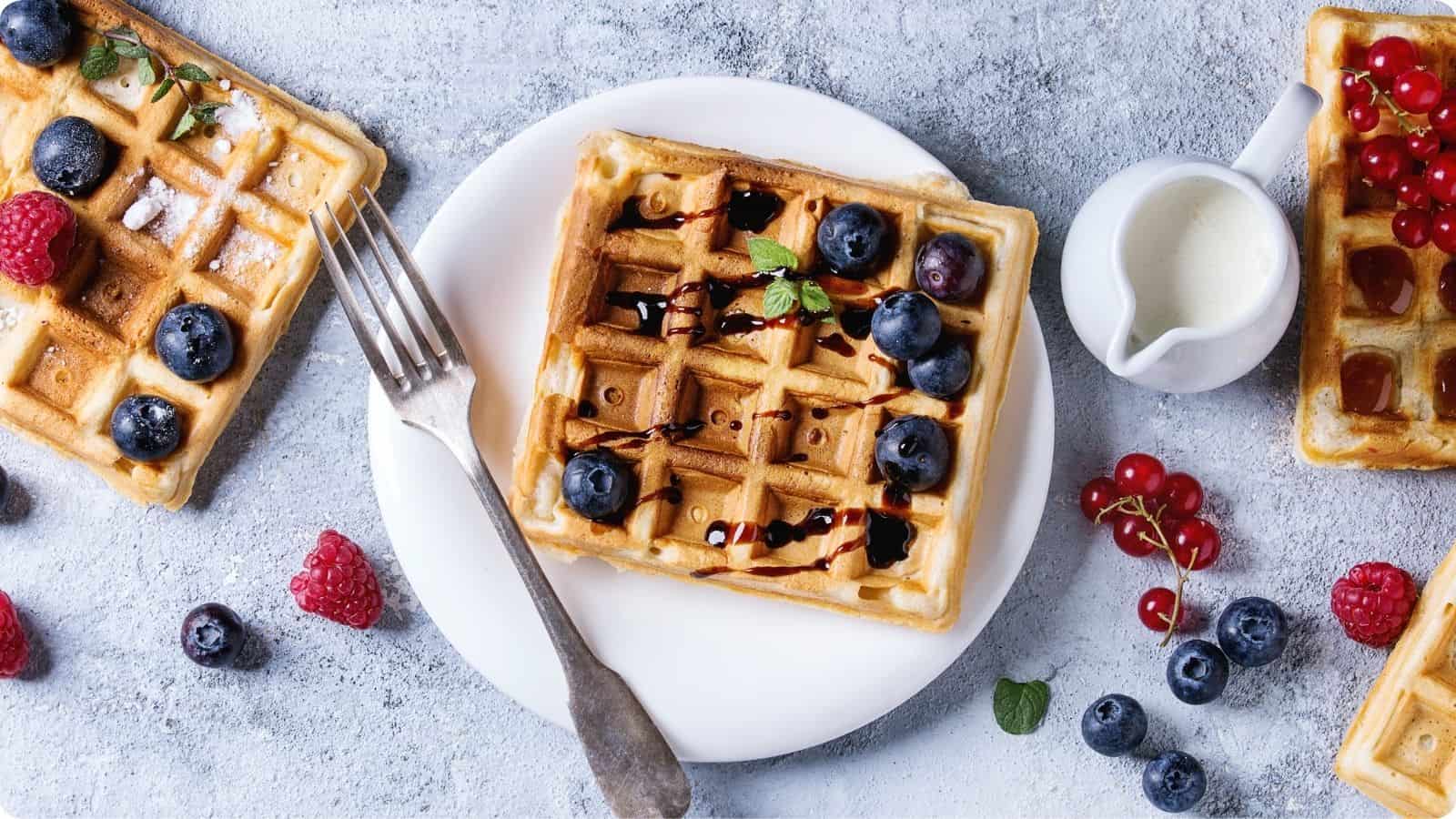 Waffles adorned with blueberries, raspberries, and cherries, drizzled with chocolate syrup and dusted with powdered sugar from a small dispenser, served on a table.⁣
⁣⁣