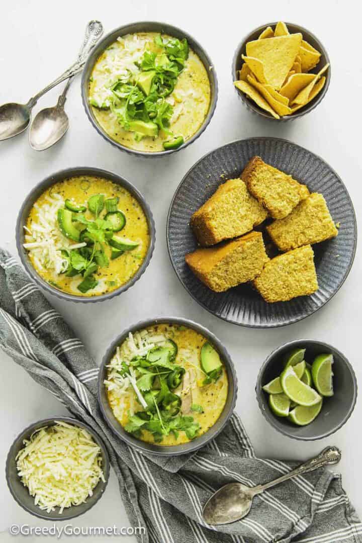Grey bowls of white chicken chili next to a plate of cornbread and bowl of cheese.